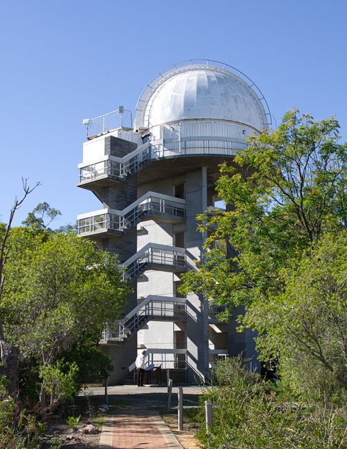 A large observatory with a white domed roof and metallic exterior, situated on a concrete structure that features an external spiral staircase wrapping around the tower. The tower is surrounded by lus